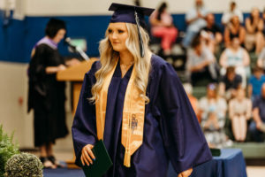 Female graduating student in cap and gown with gold scarf and commencement in the background