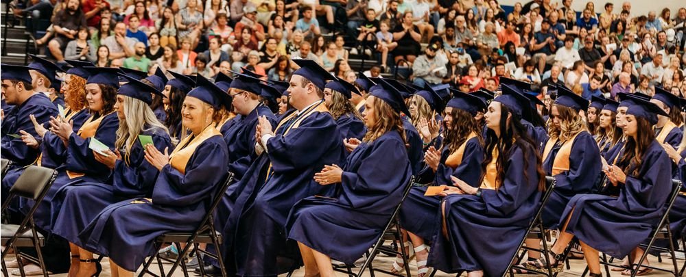 Students in caps and gowns applauding during graduation, with family audience in the background.