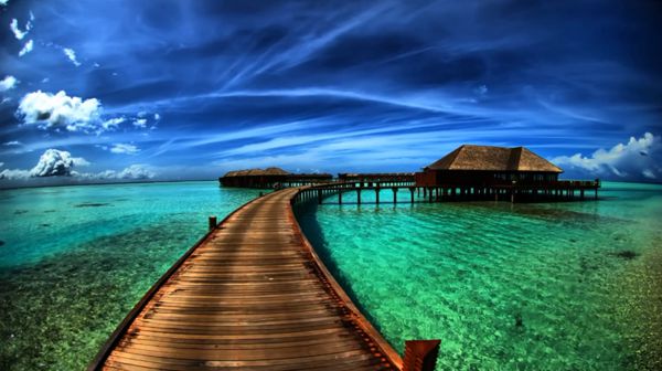 image of raised board walkway over shallow water leading to a raised hut with a cloudy sky in the background, reflected in the water.