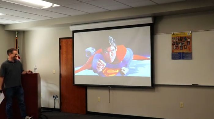 Math professor, Brian Dalpiaz giving college theme presentation, with an image on the view screen of Christopher Reeve as Superman