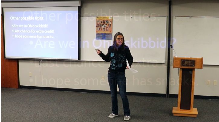 Jamie Kotewa giving College Theme Presentation, with text on a screen in the background: Other possible titles: Are we in Ohio skibbidi? Last chance for extra credit! I hope someone has snacks