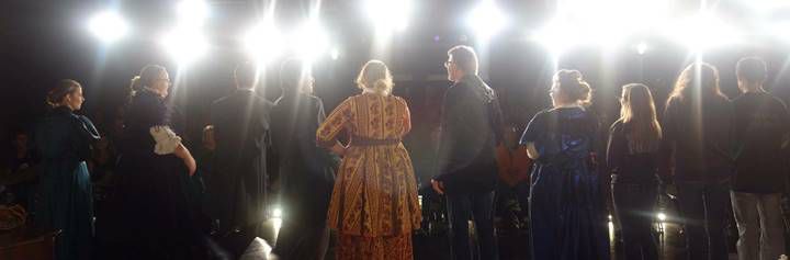 Theatre company on stage under lights, viewed from behind