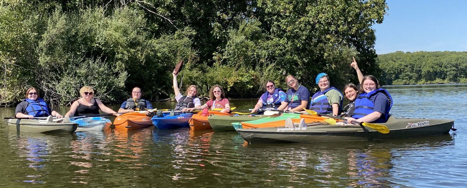 Group of ten TRIO students in canoes on a lake in front of a wooded area on the shore