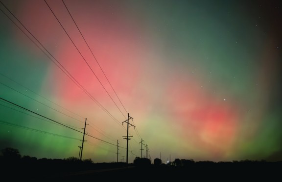 Photograph: Celestial Dance by Annalies Dowell. A nighttime photograph of a rural landscape under a sky glowing with the Northern Lights. Vivid bands of green and red aurora stretch across the dark sky above a row of electrical transmission towers silhouetted on the horizon. Stars are visible in the darker areas of sky.