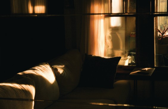 Photograph: Empty Space by Rebekkah Turner. A dimly lit living room interior. A white sofa is bathed in warm late-afternoon light streaming through gauzy curtains. The room is still and suffused with golden light and deep shadow.