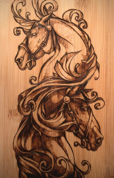 Wood burning (pyrography): The King and the Jumper by Sara Stufflebeam. Two horse heads rendered in detailed pyrography on a light bamboo or wood panel. One horse faces upward with an ornate bridle and flowing mane adorned with decorative scrollwork and curling flourishes. A second horse faces downward below it, similarly bridle-adorned. The warm brown tones of the wood burning create a rich, dimensional effect.