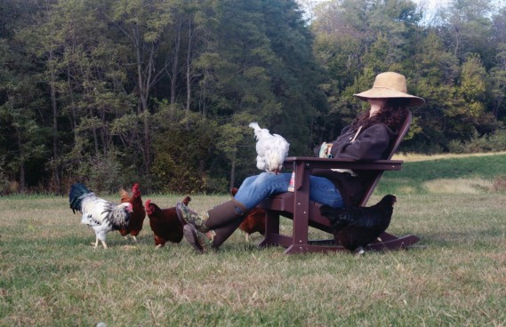 Photograph: Idle by Eliana Davis. A color photograph of a person in a wide-brimmed hat sitting in a dark red Adirondack chair in a green field. Several chickens — including a rooster and hens of various colors — roam around the chair and sit on the person's lap. A forested hillside rises behind the scene.
