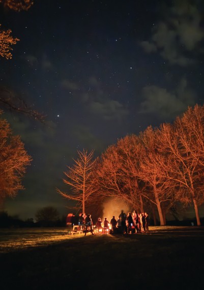 Photograph: Snapper Family by ChiChi Wang. A nighttime outdoor gathering around a bonfire. A group of silhouetted people stand around the flames beneath a star-filled dark blue sky. Bare winter trees frame the scene, their bark lit warm orange by the firelight.