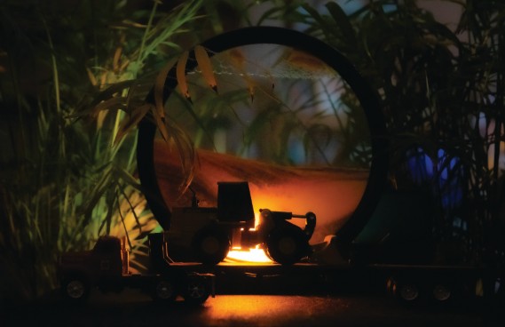 Photograph: Sunset Trailer by Rebekkah Turner. A creative still-life or diorama photograph showing a miniature locomotive or trailer scene viewed through a large circular opening, with warm amber firelight glowing at the center against a backdrop of lush green tropical foliage and deep blue tones.